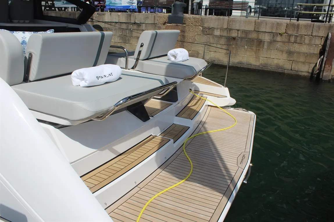 Luxury boat deck view featuring two gray cushioned seats with rolled towels labeled "FAIRLINE" and yellow hose on wooden flooring.